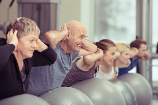 Happy elderly couple exercising in a pilates class at the gym with three other younger people toning and strengthening their muscles using gym balls, focus to the senior man and woman Happy elderly couple exercising in a pilates class at the gym with three other younger people toning and strengthening their muscles using gym balls, focus to the senior man and woman